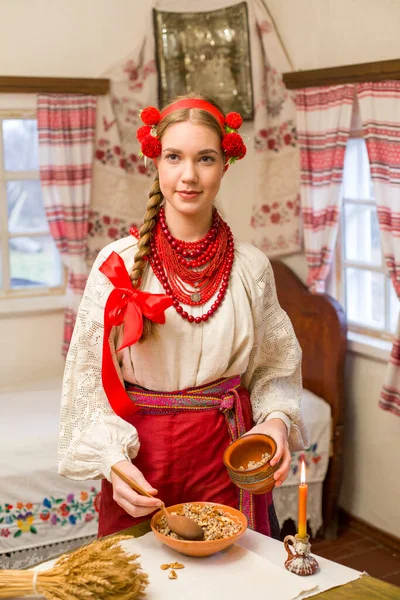 Menina bonita em vestido nacional está preparando um jantar festivo. Em uma grinalda bonita e um vestido bordado vermelho. Celebração familiar e celebração do costume nacional. Tigela com kutia - tradicional — Fotografia de Stock