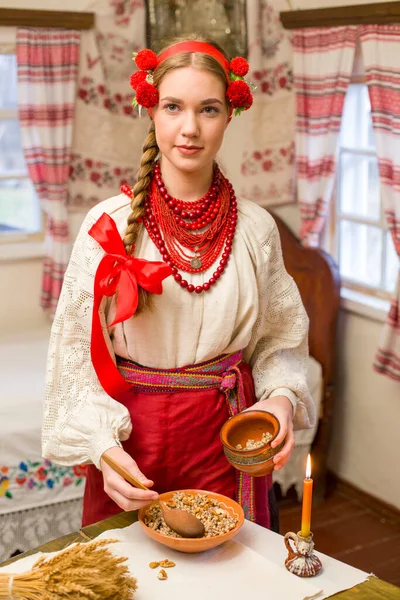 Piękna dziewczyna w sukience narodowej przygotowuje uroczysty obiad. W pięknym wieńcu i czerwonej haftowanej sukience. Rodzinne świętowanie i świętowanie narodowego zwyczaju. Miska z kutią - tradycyjna — Zdjęcie stockowe