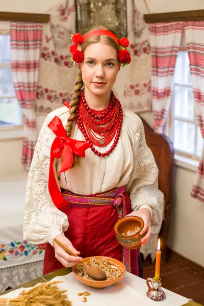Beautiful girl in national dress is preparing a festive dinner. In a beautiful wreath and a red embroidered dress. Family celebration and celebration of national custom. Bowl with kutia - traditional — Stock Photo, Image