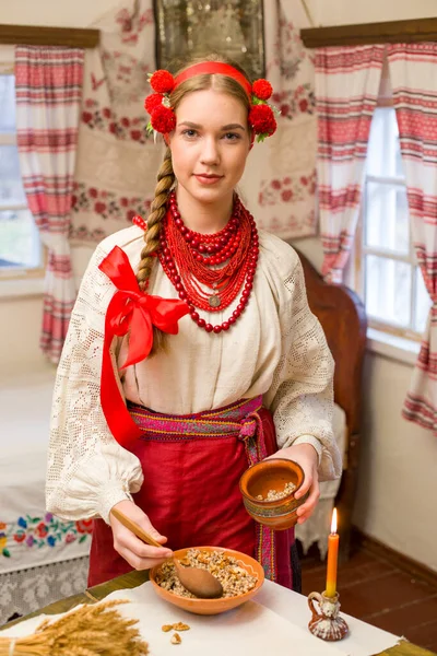 Menina bonita em vestido nacional está preparando um jantar festivo. Em uma grinalda bonita e um vestido bordado vermelho. Celebração familiar e celebração do costume nacional. Tigela com kutia - tradicional — Fotografia de Stock