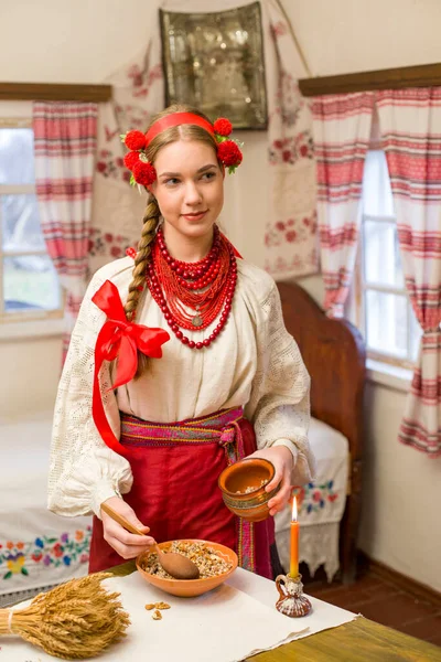 穿着民族服装的漂亮姑娘正在准备喜庆的晚餐.戴着美丽的花环和红色绣花裙。家庭庆祝和民族风俗的庆祝。与kutia -传统的碗 — 图库照片