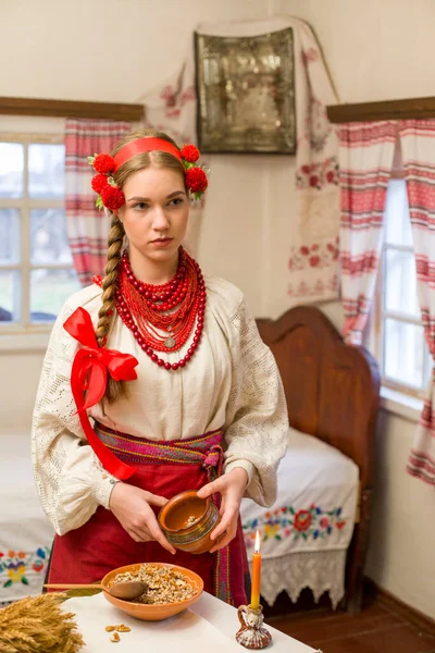 Menina bonita em vestido nacional está preparando um jantar festivo. Em uma grinalda bonita e um vestido bordado vermelho. Celebração familiar e celebração do costume nacional. Tigela com kutia - tradicional — Fotografia de Stock