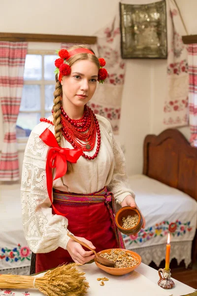 Όμορφο κορίτσι με εθνικό φόρεμα ετοιμάζει ένα εορταστικό δείπνο. Με ένα όμορφο στεφάνι και ένα κόκκινο κεντημένο φόρεμα. Οικογενειακή γιορτή και εορτασμός των εθνικών εθίμων. Μπολ με kutia - παραδοσιακό — Φωτογραφία Αρχείου
