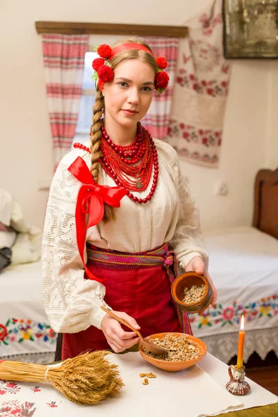 Menina bonita em vestido nacional está preparando um jantar festivo. Em uma grinalda bonita e um vestido bordado vermelho. Celebração familiar e celebração do costume nacional. Tigela com kutia - tradicional — Fotografia de Stock