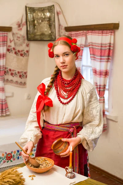 Hermosa chica vestida con ropa nacional está preparando una cena festiva. En una hermosa corona y un vestido rojo bordado. Celebración familiar y celebración de la costumbre nacional. Bollo con kutia - Tradicional — Foto de Stock