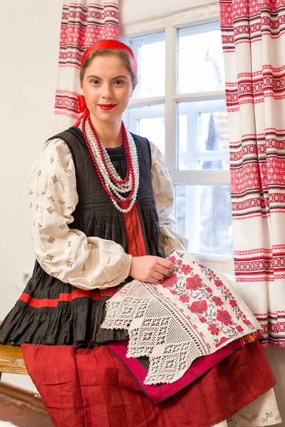 Menina bonita em um traje nacional do século 19. Embroiders um belo padrão perto da janela. Com uma grinalda e fitas. Vila, campo na foto. Em um lenço de cabeça. — Fotografia de Stock