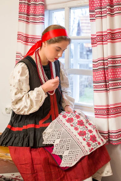Menina bonita em um traje nacional do século 19. Embroiders um belo padrão perto da janela. Com uma grinalda e fitas. Vila, campo na foto. Em um lenço de cabeça. — Fotografia de Stock