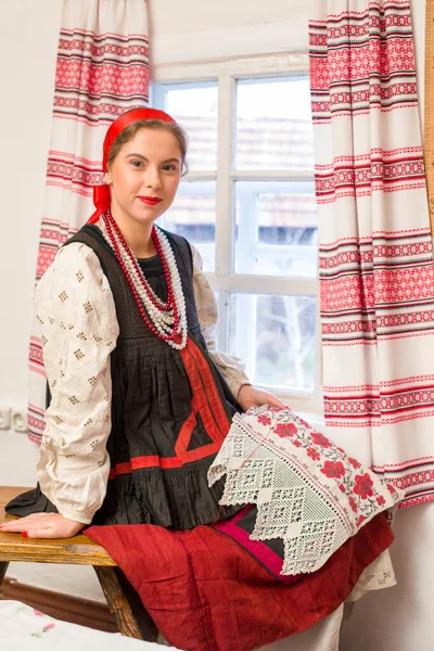 Beautiful young girl in a national costume of the 19th century. Embroiders a beautiful pattern near the window. With a wreath and ribbons. Village, countryside in the photo. In a head scarf. — Stock Photo, Image