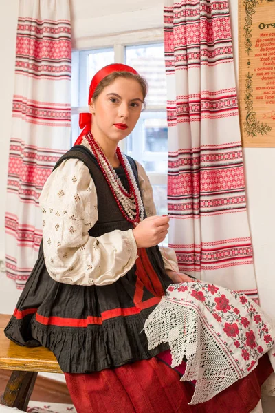 Menina bonita em um traje nacional do século 19. Embroiders um belo padrão perto da janela. Com uma grinalda e fitas. Vila, campo na foto. Em um lenço de cabeça. — Fotografia de Stock