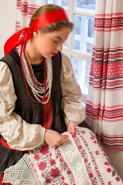 Menina bonita em um traje nacional do século 19. Embroiders um belo padrão perto da janela. Com uma grinalda e fitas. Vila, campo na foto. Em um lenço de cabeça. — Fotografia de Stock