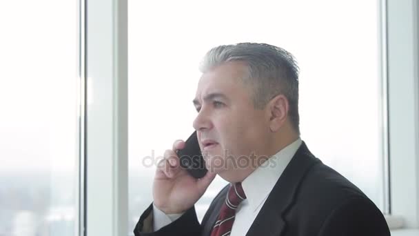 Üzletember egy öltöny öltöny és beszél a telefonon közelében a panoráma ablakokkal. Közelről. Ő volt nagyon riasztónak tartja.