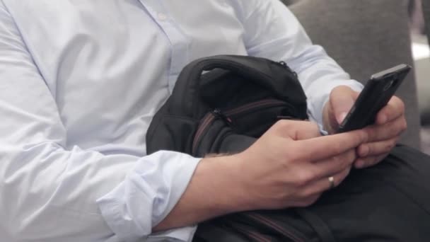Um homem sentado em uma cadeira de camisa está digitando algo no telefone — Vídeo de Stock