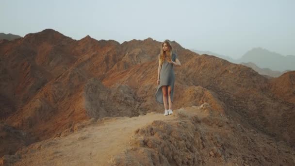 Joven mujer rubia de pie en la cima de la montaña. Paisaje de montaña con puesta de sol, cámara lenta, 4k — Vídeos de Stock