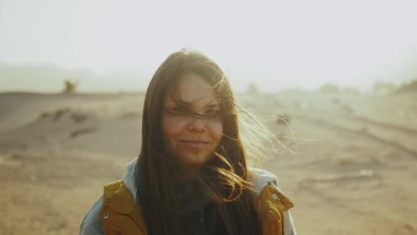 Portret van een mooie vrouw bij zonsondergang. Gelukkige jonge vrouw staande in de wind tegen een woestijn zonsondergang landschap, slow motion. — Stockvideo