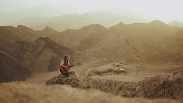 Kvinna spelar gitarr och sjunger i ökenlandskap, öken berg bakgrund, slow motion, 4k — Stockvideo