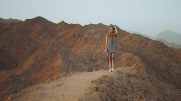 Joven mujer rubia de pie en la cima de la montaña. Paisaje de montaña con puesta de sol, cámara lenta, 4k — Vídeos de Stock