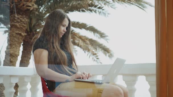 Krásná studentka nebo na volné noze pracující na notebooku, žena sedící na balkóně s výhledem shora na dlaň a moře, rychlé video, zpomalení, 4k — Stock video
