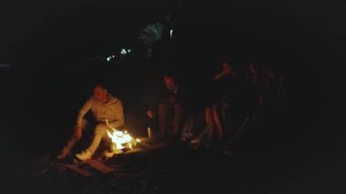 Bir grup arkadaş, gece deniz kenarındaki kumsalda kamp ateşinin yanında eğleniyor. Hızlı video, ağır çekim, 4k