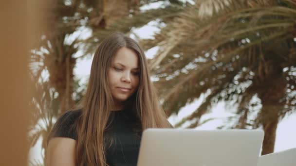 Hermosa estudiante o freelancer que trabaja en un ordenador portátil, mujer sentada en el balcón con vista superior de la palma y el mar, vídeo rápido, cámara lenta, 4k — Vídeos de Stock