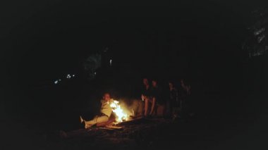 Bir grup arkadaş, gece deniz kenarındaki kumsalda kamp ateşinin yanında eğleniyor. Hızlı video, ağır çekim, 4k