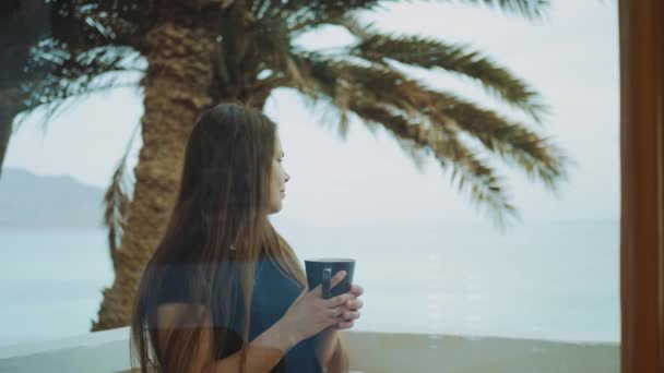 Alleen vrouw is het drinken van koffie of thee in de ochtend op het balkon met blauwe uitzicht op zee, standpunt vanuit het raam, snelle video, slow motion, 4k — Stockvideo