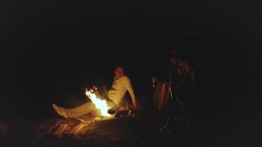 Bir grup arkadaş, gece deniz kenarındaki kumsalda kamp ateşinin yanında eğleniyor. Hızlı video, ağır çekim, 4k