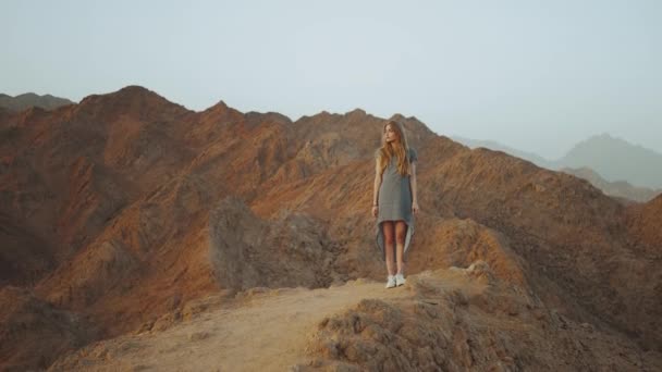 Joven mujer rubia de pie en la cima de la montaña. Paisaje de montaña con puesta de sol, cámara lenta, 4k — Vídeos de Stock