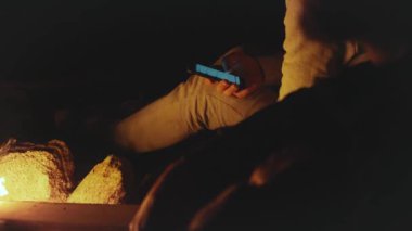 Bir grup arkadaş, gece deniz kenarındaki kumsalda kamp ateşinin yanında eğleniyorlar. Akıllı telefon tutan adam, hızlı video, ağır çekim, 4k.