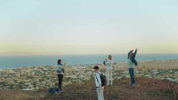 Skupina přátel relaxovat na vrcholu hory při západu slunce a užívat si výhled na moře - přátelství, mládež, zpomalení, 4k — Stock video