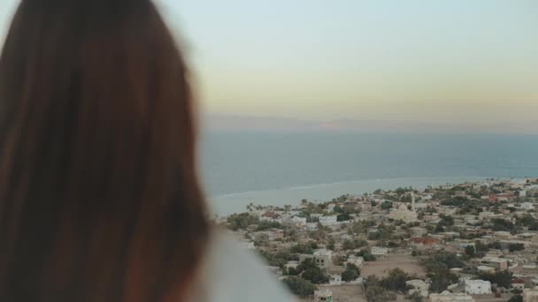 Vue de la mer bleue et petit village de dos de femme. Fille rester au sommet de la montagne et regarder dans l'horizon, au ralenti, 4k — Video