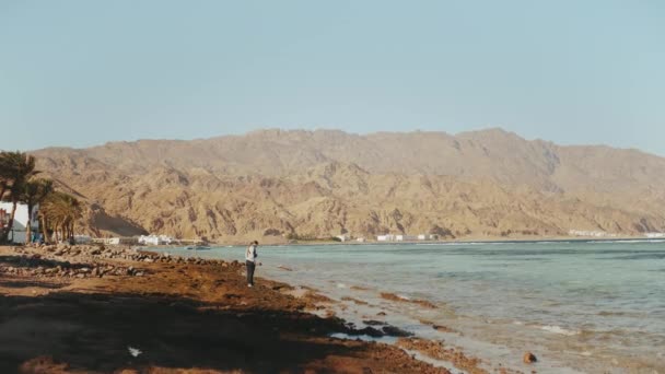 Mujer bonita caminando sola en la playa costera pedregosa cerca del mar, las olas se rompen en la orilla, Egipto Sinaí montaña en el fondo, cámara lenta, 4k — Vídeo de stock
