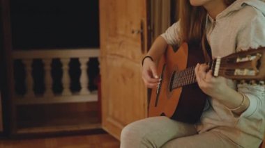 Gözlüklü sarışın kız gitar çalıyor ve rahat bir dairede sandalyede şarkı söylüyor. Bir kadın içeride müzik yaratır, ağır çekim, 4k