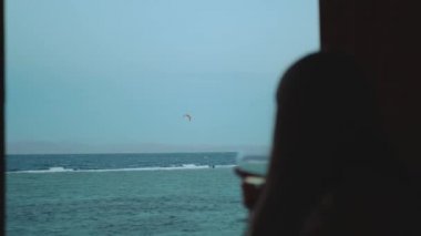 Kadın şarap içiyor ve bak insanlar Dahab Mısır 'da temiz suda nasıl sörf yapıyor. Arka planda dağlar olan mavi denizi keşfetmek ve insanlar uçurtma sörfü, yavaş çekim, 4k