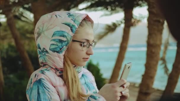 Mulher loira jovem em óculos tirar foto por telefone, vento forte, mar e palmas no fundo, câmera lenta, 4k — Vídeo de Stock