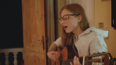 Gözlüklü sarışın kız gitar çalıyor ve rahat bir dairede sandalyede şarkı söylüyor. Bir kadın içeride müzik yaratır, ağır çekim, 4k