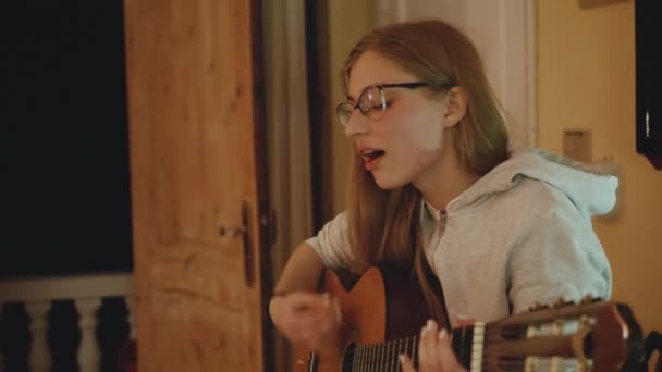 La chica rubia en gafas toca la guitarra y canta en la silla en un acogedor apartamento. Una mujer crea música en el interior, cámara lenta, 4k — Vídeos de Stock