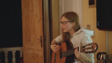 Gözlüklü sarışın kız gitar çalıyor ve rahat bir dairede sandalyede şarkı söylüyor. Bir kadın içeride müzik yaratır, ağır çekim, 4k