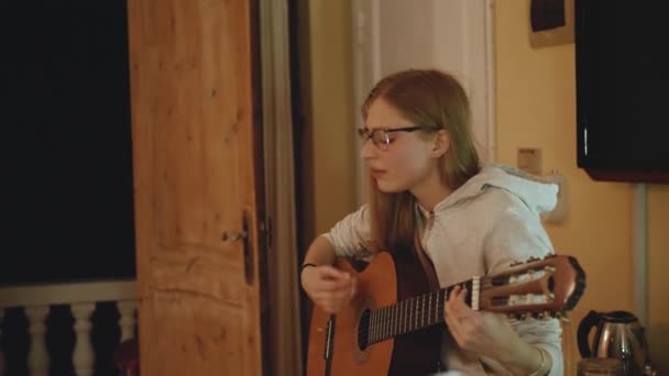 A menina loira de óculos toca guitarra e canta na cadeira em apartamento acolhedor. Uma mulher cria música dentro de casa, câmera lenta, 4k — Vídeo de Stock