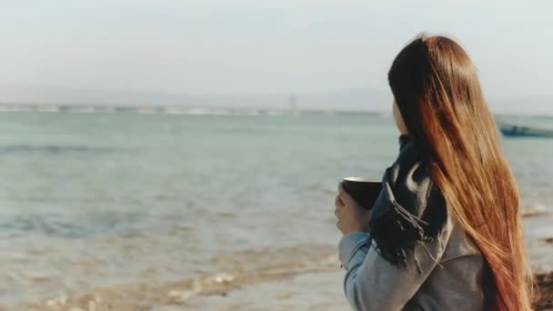 Жінка п'є каву або чай на пляжі з синім видом на море, жінка дивиться на горизонт, швидке відео, повільний рух, 4k — стокове відео