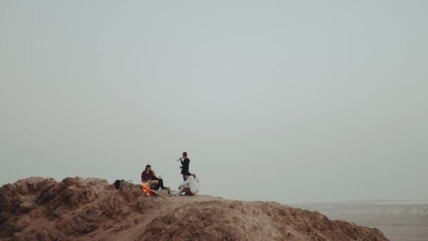 Groep mensen ontspannen op de top van een berg bij zonsondergang, zitten in de buurt van vreugdevuur, genieten van hun tijd - vriendschap, jeugd, slow motion, full hd — Stockvideo