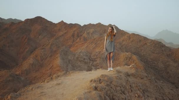 Eine junge blonde Frau im langen Kleid geht auf den Gipfel des Berges. Berglandschaft mit Sonnenuntergang, Zeitlupe, voller — Stockvideo