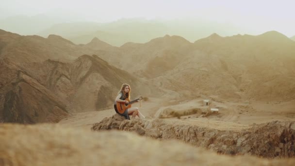 Kvinna som spelar gitarr och sjunger i ökenlandskap, öken berg bakgrund, slow motion, full hd — Stockvideo