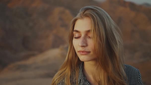 노을에 눈을 감고 있는 예쁜 금발 여자의 초상화. 사막의 산에 서 있는 행복 한 젊은 여성, 일몰 풍경, 느린 움직임, 꽉 찬 Hd — 비디오