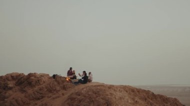 Gün batımında bir dağın tepesinde dinlenen bir grup insan, şenlik ateşinin yanında oturmuş, eğleniyorlar - arkadaşlık, gençlik, ağır çekim, tam kapasite