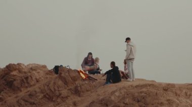 Gün batımında bir dağın tepesinde dinlenen bir grup insan, şenlik ateşinin yanında oturmuş, eğleniyorlar - arkadaşlık, gençlik, ağır çekim, tam kapasite