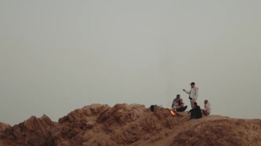 Gün batımında bir dağın tepesinde dinlenen bir grup insan, şenlik ateşinin yanında oturmuş, eğleniyorlar - arkadaşlık, gençlik, ağır çekim, tam kapasite