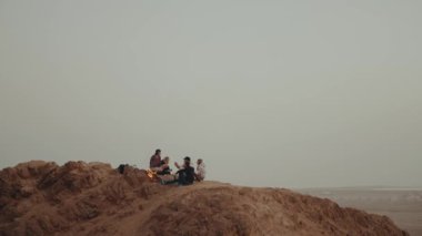 Gün batımında bir dağın tepesinde dinlenen bir grup insan, şenlik ateşinin yanında oturmuş, eğleniyorlar - arkadaşlık, gençlik, ağır çekim, tam kapasite