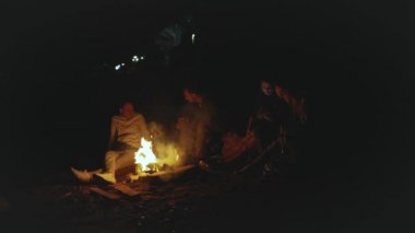 Bir grup arkadaş, gece deniz kenarındaki kumsalda kamp ateşinin yanında eğleniyor. Hızlı video, yavaş çekim, tam yol ileri.