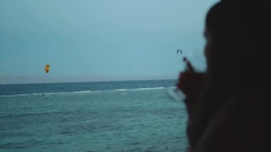 Kadın şarap içiyor ve bak insanlar Dahab Mısır 'da temiz suda nasıl sörf yapıyor. Arka planda dağlarla mavi denizi keşfetmek ve insanlar uçurtma sörfü, yavaş çekim, tam yol ileri.