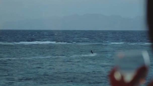 Жінка п'є вино і дивиться, як люди кидають серфінг у чудовій чистій воді в Дахабі. Дослідження синього моря з горами на задньому плані і люди змітають серфінг, повільний рух, повний hd — стокове відео